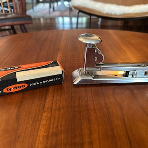 Puede incluir: Una grapadora de metal cromado con un mecanismo de émbolo superior redondeado, colocada junto a una caja de cartón rectangular etiquetada como Ty-Dee Brand con el texto impreso a ty-dee job's a better job en colores naranja, negro y blanco.
