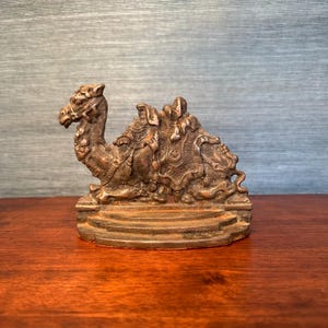 May include: A detailed bronze camel figurine, resting on a tiered base. The camel is adorned with intricate saddle details and bridle. The sculpture is a decorative item, likely for display.