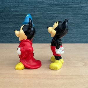 May include: Two Mickey Mouse figurines. One is dressed as a sorcerer in a red robe and blue hat with a gold star. The other Mickey Mouse is in a black shirt, red shorts, and yellow shoes. Both have white gloves.
