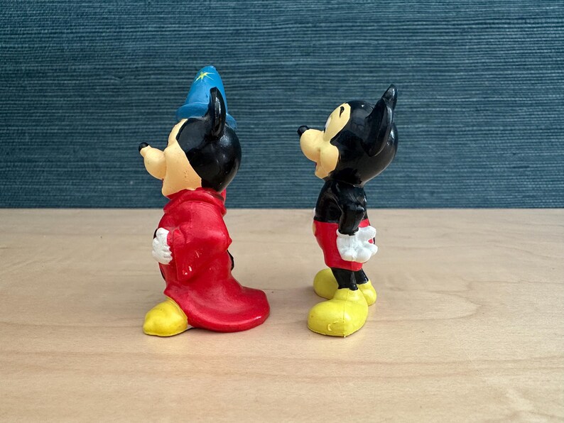 May include: Two Mickey Mouse figurines. One is dressed as a sorcerer in a red robe and blue hat with a gold star. The other Mickey Mouse is in a black shirt, red shorts, and yellow shoes. Both have white gloves.