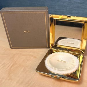 Puede incluir: Un estuche compacto dorado con espejo y borla, junto a una caja marrón. El estuche compacto está abierto, revelando una borla blanca con la palabra "Avon" impresa. La caja también tiene la palabra "Avon" impresa.