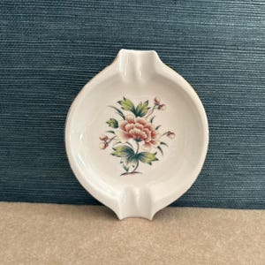 May include: White ceramic ashtray with a floral design. The ashtray has a rounded shape with a small lip and a decorative floral pattern in the center, featuring red, green, and pink hues. The rim has a gold trim.