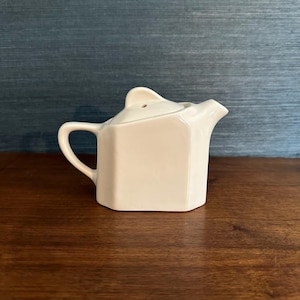Op de afbeelding: Kleine, crèmekleurige keramische theepot met een deksel en een gebogen handvat. De theepot heeft een tuit en een achthoekige vorm. Hij staat op een donkerhouten oppervlak tegen een blauwe, getextureerde achtergrond.