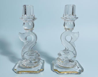 May include: Two clear glass candle holders with a dolphin design. The candle holders have a square base with gold trim and a dolphin-shaped stem. The top of the candle holders are designed to hold a candle.