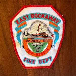 Könnte beinhalten: Ein bestickter Aufnäher mit rotem Rand und der Aufschrift EAST ROCKAWAY FIRE DEPT. Das Design zeigt ein Boot, ein grünes Dreieck mit Fisch und Krabbe sowie ein orangefarbenes Banner mit der Aufschrift ORGANIZED 1893 auf weißem Grund.