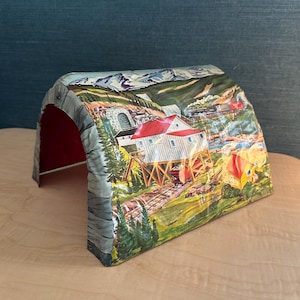 May include: A vintage toy train tunnel with a detailed landscape scene. The tunnel entrance is gray, with a red interior. The painted scene depicts a mountain range, a building with a red roof, and a train on a bridge.