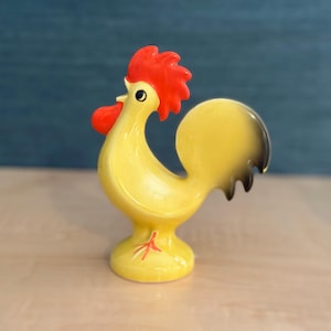 May include: A ceramic rooster figurine featuring a bright yellow body, a vibrant red comb and wattle, and a black-tipped tail. The rooster stands on a circular base with painted red feet, showcasing a smooth, glossy finish and a stylized design.