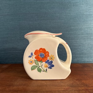 May include: White ceramic jug with a red rim and floral design. The jug has a handle and a lid. The flowers are orange, blue, and yellow.