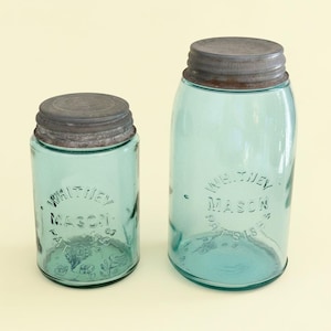 Könnte beinhalten: Zwei Vintage-Gläser in Blaugrün mit Metalldeckeln. Die Gläser sind mit den Worten "Whitney Mason" und dekorativen Elementen geprägt. Ein Glas ist höher als das andere. Die Deckel sind dunkelgrau.