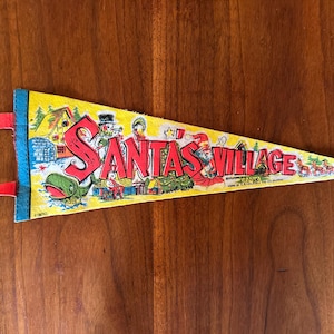May include: A yellow triangular felt pennant featuring the red text Santa's Village. The graphic includes a green whale, a snowman, a house, and a reindeer sleigh. It has a blue vertical strip on the left side with two red felt ties attached.
