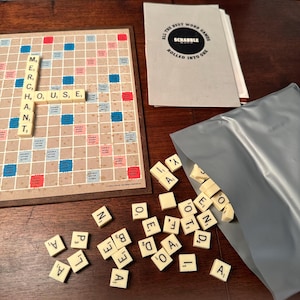 Pode incluir: Um jogo de tabuleiro Scrabble sobre uma superfície de madeira. O tabuleiro exibe as palavras "MERCHANT" e "HOUSE". Peças espalhadas e uma bolsa cinza com mais peças são visíveis. A caixa do jogo diz "ALL THE BEST WORD GAMES SCRABBLE ROLLED INTO ONE".