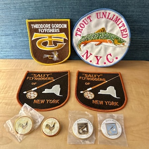 Może przedstawiać: Kolekcja naszywek i przypinek o tematyce wędkarskiej. Na naszywkach znajdują się napisy takie jak "Theodore Gordon Flyfishers" i "Salty Flyrodders of New York". Przypinki przedstawiają koguta i wzory rocznicowe. Przedmioty znajdują się na drewnianej powierzchni.