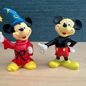 May include: Two Mickey Mouse figurines. One is dressed as a sorcerer in a red robe and blue hat, holding a star-topped wand. The other Mickey Mouse figure is in a black shirt, red shorts, and yellow shoes. Both figures have black ears and white gloves.