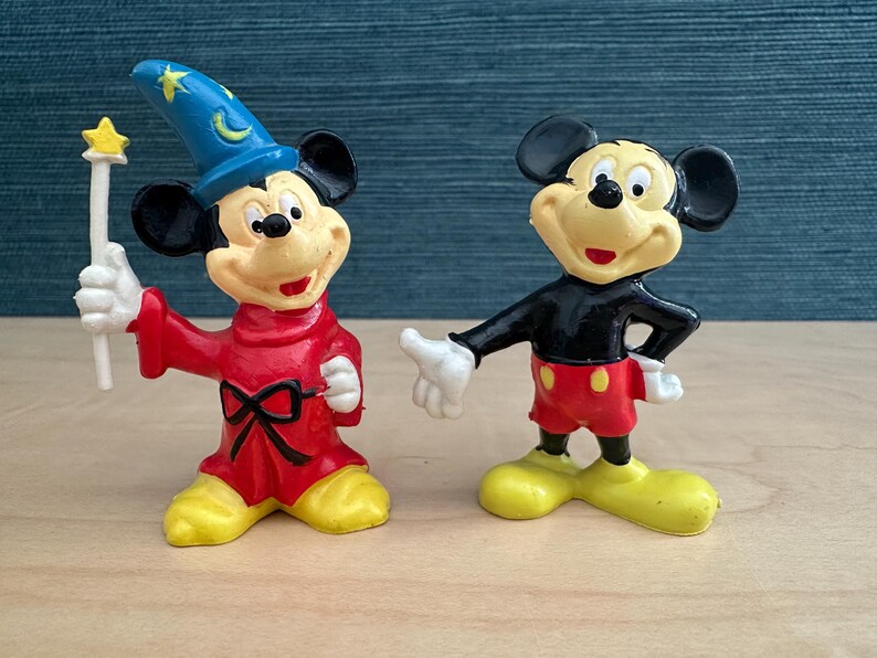 May include: Two Mickey Mouse figurines. One is dressed as a sorcerer in a red robe and blue hat, holding a star-topped wand. The other Mickey Mouse figure is in a black shirt, red shorts, and yellow shoes. Both figures have black ears and white gloves.
