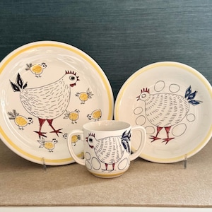 Puede incluir: Un juego de vajilla de cerámica con un diseño de gallina y pollitos. El juego incluye un plato grande, un plato más pequeño y una taza con asas. Los platos tienen un borde amarillo y las ilustraciones de gallinas son en negro, rojo y amarillo.