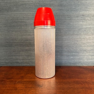 May include: A tall, cylindrical container with a bright red cap. The body of the container is clear with vertical ribbed detailing. The container is sitting on a dark wooden surface.