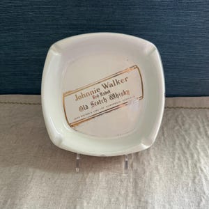 Pode incluir: Cinzeiro quadrado branco com cantos arredondados. O cinzeiro apresenta o texto "Johnnie Walker Red Label Old Scotch Whisky" em letras douradas e vermelhas. O cinzeiro &eacute; feito de cer&acirc;mica ou porcelana.