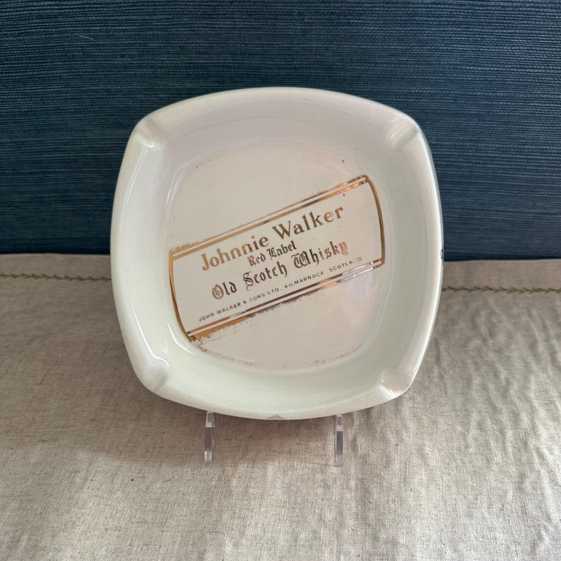 Pode incluir: Cinzeiro quadrado branco com cantos arredondados. O cinzeiro apresenta o texto "Johnnie Walker Red Label Old Scotch Whisky" em letras douradas e vermelhas. O cinzeiro &eacute; feito de cer&acirc;mica ou porcelana.