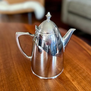 May include: A polished silver-tone metal teapot featuring a tapered cylindrical body, a hinged domed lid with a small pointed finial, a curved spout, and an angular handle. The reflective surface shows light highlights and surrounding environment.