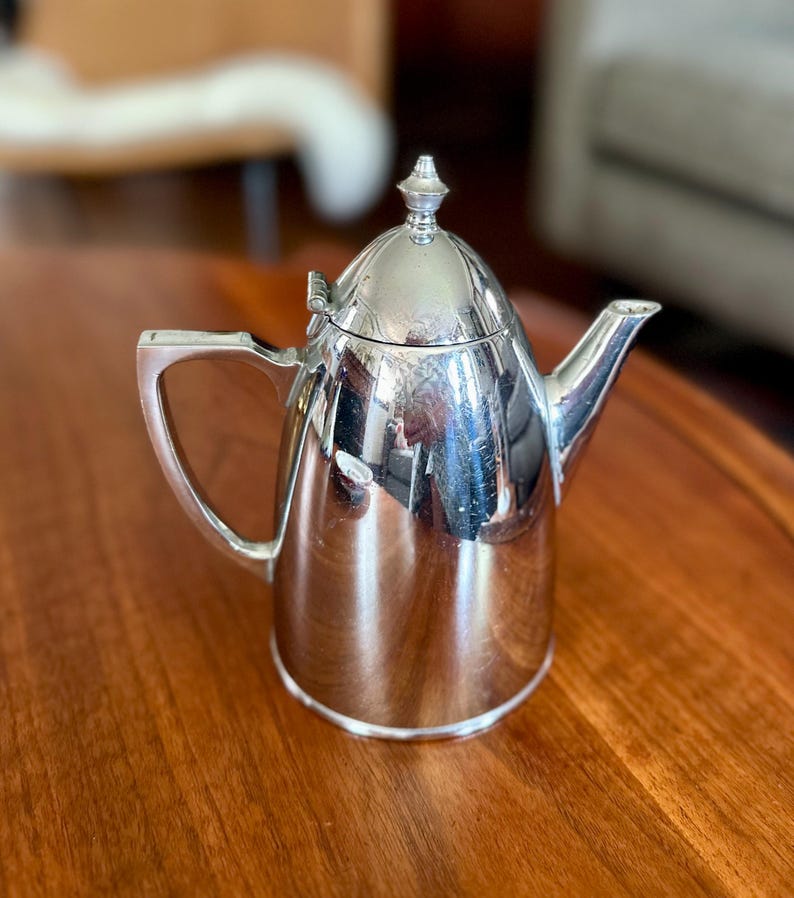 May include: A polished silver-tone metal teapot featuring a tapered cylindrical body, a hinged domed lid with a small pointed finial, a curved spout, and an angular handle. The reflective surface shows light highlights and surrounding environment.
