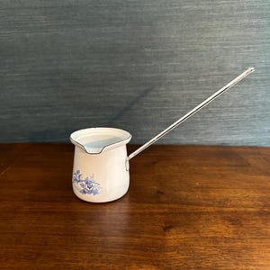 Puede incluir: Una jarra de esmalte blanco con un asa larga y un diseño floral azul. La jarra tiene un cuerpo redondeado y un pico. El asa es fina y se extiende desde el lateral. La jarra está sobre una superficie de madera.