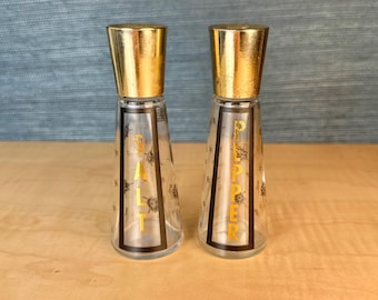 May include: A pair of vintage salt and pepper shakers. The clear glass shakers have gold-colored lids and black and gold accents. The word "SALT" and "PEPPER" are printed in gold on the front of each shaker. The shakers are on a wooden surface.