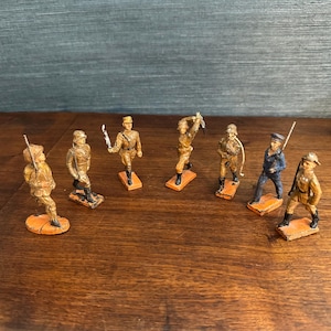 May include: Eight vintage toy soldiers in various poses. The figures are painted in shades of tan, brown, and blue. Each soldier stands on a small, orange rectangular base. The toys are arranged on a wooden surface.