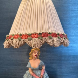 Puede incluir: Una l&aacute;mpara de mesa vintage con una pantalla de l&aacute;mpara blanca plisada adornada con una cinta verde y roja. La base de la l&aacute;mpara presenta una figurita de porcelana de una mujer con un vestido azul que sostiene un perro blanco.