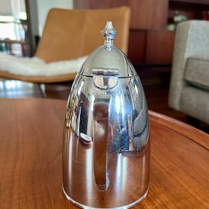 May include: A polished silver-tone metal thermal carafe with a rounded, dome-shaped lid featuring a small decorative finial at the top. The carafe has a smooth, reflective surface, a vertical handle on the side, and a hinged lid mechanism.