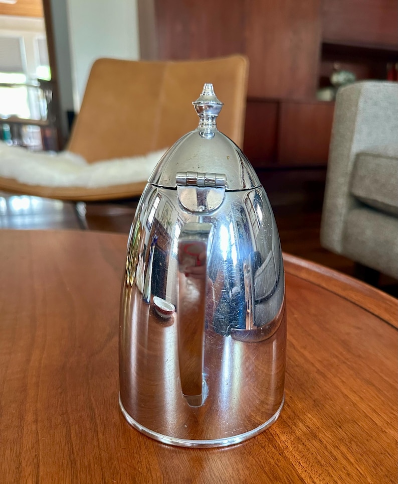 May include: A polished silver-tone metal thermal carafe with a rounded, dome-shaped lid featuring a small decorative finial at the top. The carafe has a smooth, reflective surface, a vertical handle on the side, and a hinged lid mechanism.