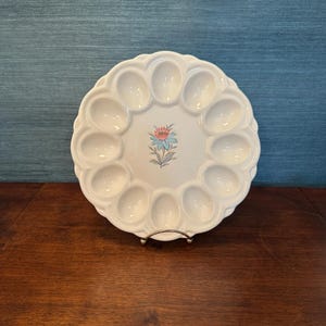 May include: White ceramic egg serving platter with 12 egg-shaped indentations around the edge. A floral design in pink and blue is in the center. The platter is on a silver metal stand.