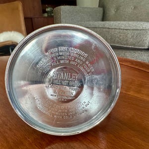 May include: The circular silver-colored metal base of a Stanley vacuum bottle features embossed text including instructions for hot and cold liquids, the brand name Stanley, the phrase It Will Not Break, and Landers, Frary & Clark, New Britain, Conn. U.S.A.