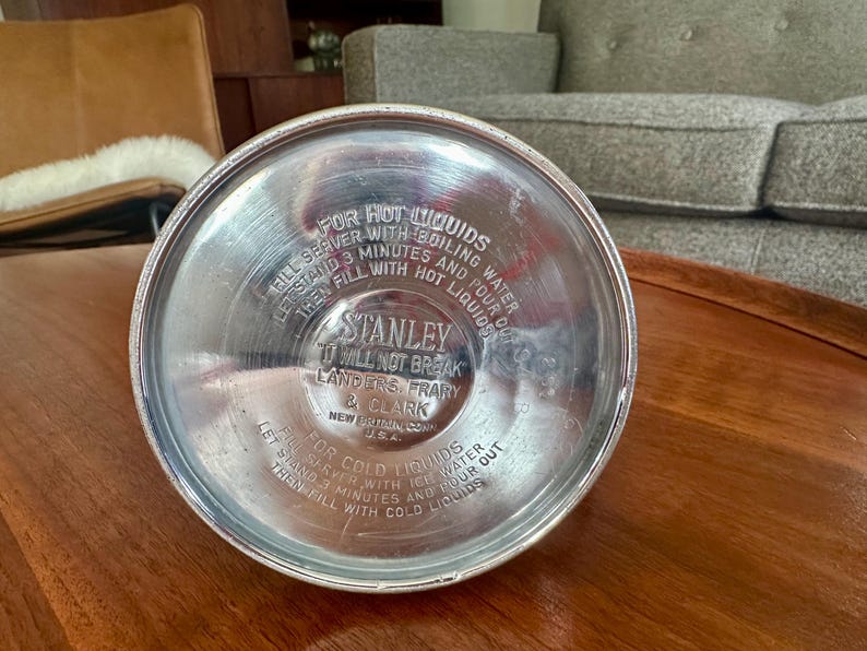 May include: The circular silver-colored metal base of a Stanley vacuum bottle features embossed text including instructions for hot and cold liquids, the brand name Stanley, the phrase It Will Not Break, and Landers, Frary & Clark, New Britain, Conn. U.S.A.