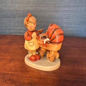 May include: A ceramic figurine of a young girl in a red dress and yellow apron, standing next to a baby carriage. The carriage has an orange canopy and wooden frame. The figurine is on a white oval base.