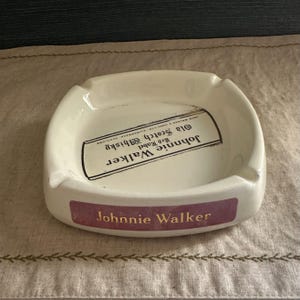 Pode incluir: Cinzeiro de cer&acirc;mica creme com o logotipo "Johnnie Walker" em letras douradas em uma faixa bord&ocirc;. O cinzeiro apresenta um r&oacute;tulo retangular com "Johnnie Walker Red Label Old Scotch Whisky" impresso em preto.