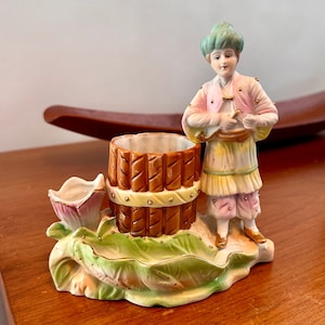 Könnte beinhalten: Eine Porzellanfigur eines Menschen in einem grünen Turban und rosa und gelber Kleidung, die neben einem braunen Fass steht. Die Figur steht auf einem grünen und weißen Blatt.