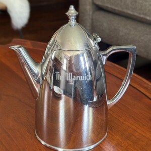 May include: A polished silver-toned metal teapot with a hinged lid, a pointed finial, and a curved handle. The side of the teapot features the engraved text The Warwick. The surface is reflective, showing light highlights and subtle surface scratches.