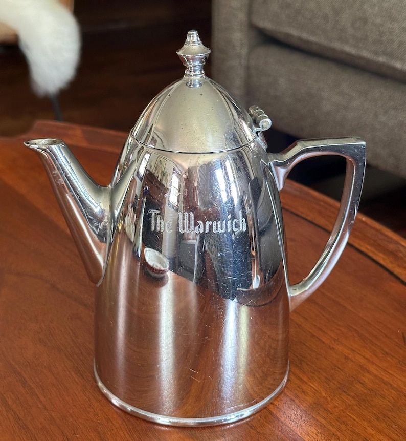 May include: A polished silver-toned metal teapot with a hinged lid, a pointed finial, and a curved handle. The side of the teapot features the engraved text The Warwick. The surface is reflective, showing light highlights and subtle surface scratches.