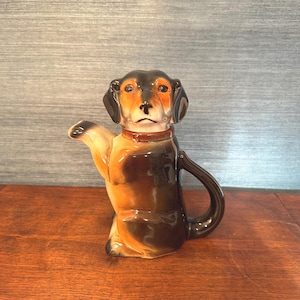 Pode incluir: Bule de cerâmica em forma de dachshund, com esquema de cores castanho, preto e bege. O bule tem uma pega e um bico, e usa uma coleira vermelha. O cão está posicionado como se estivesse sentado e a acenar.
