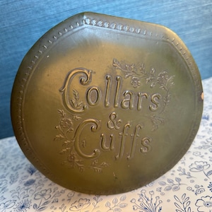 May include: An antique, olive-green metal container with the embossed text "Collars & Cuffs" in an elegant, vintage font. The container has a rounded shape with a decorative floral border.