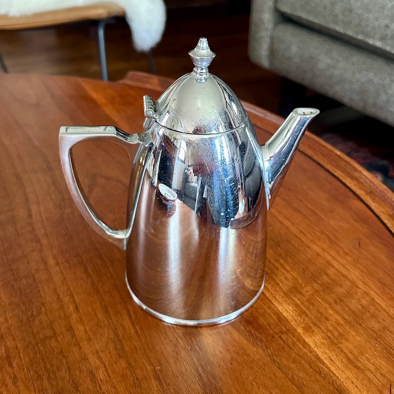 May include: A polished silver-tone metal teapot featuring a conical lid with a small finial, a curved spout, and a flat-sided handle. The reflective surface shows distorted surroundings, and the body has a tapered cylindrical shape.