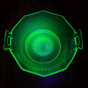 May include: A glowing green glass serving bowl with a geometric design. The bowl has a central circular pattern and a radiating design on the inside. It features handles on either side and a textured exterior.