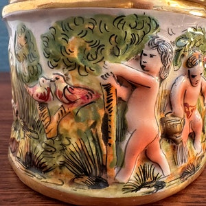 Puede incluir: Un tarro de cer&aacute;mica con un borde dorado y un dise&ntilde;o pintado a mano. El dise&ntilde;o presenta dos p&aacute;jaros, dos querubines y una exuberante escena de bosque verde.