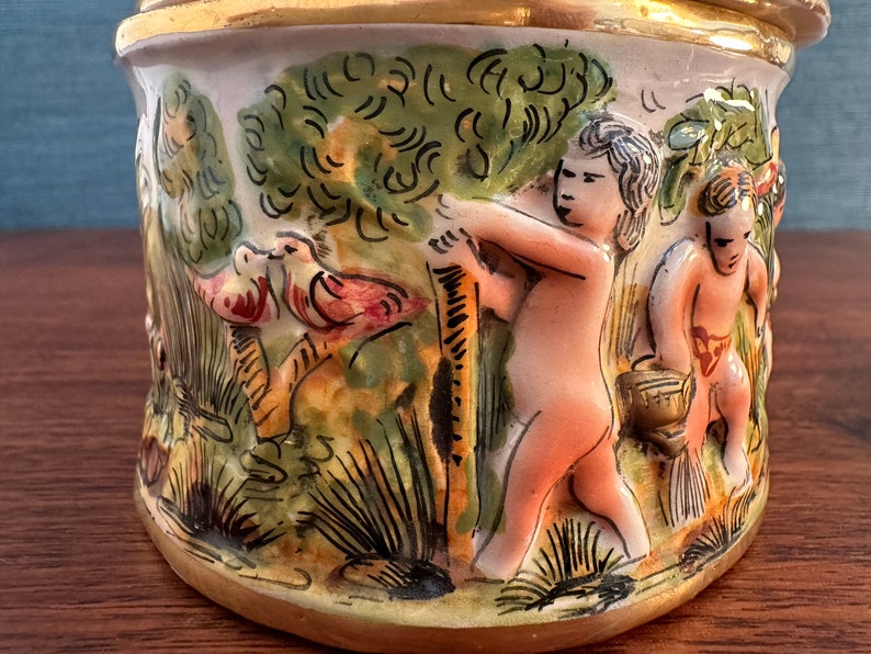 Puede incluir: Un tarro de cer&aacute;mica con un borde dorado y un dise&ntilde;o pintado a mano. El dise&ntilde;o presenta dos p&aacute;jaros, dos querubines y una exuberante escena de bosque verde.