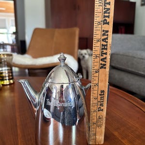 May include: A polished silver-tone metal teapot with a tapered body, a hinged lid, and a spout. The side of the teapot features the engraved text 'The Warwick'. A wooden ruler is positioned vertically next to it, showing a height of 6 inches.