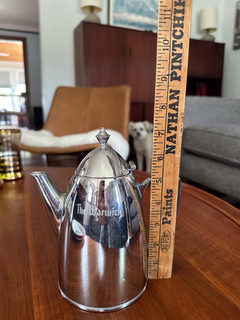 May include: A polished silver-tone metal teapot with a tapered body, a hinged lid, and a spout. The side of the teapot features the engraved text 'The Warwick'. A wooden ruler is positioned vertically next to it, showing a height of 6 inches.