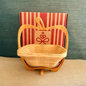 May include: A natural bamboo apple-shaped basket with a folding design. The basket has a handle and a base for support. The background features a box with red and white stripes and a logo.