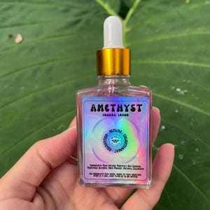 May include: A small glass bottle with a gold cap and dropper. The bottle is filled with a clear liquid and has a holographic label that says "Amethyst Chakra: Crown" and features a circle with the words "Relaxation, Healing, Enlightenment".