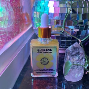 May include: A clear glass bottle with a gold cap and dropper filled with a yellow liquid. The bottle label reads "Citrine Chakra: Solar Plexus" and features a yellow flower with the words "Manifestation, Confidence, Joy" inside.