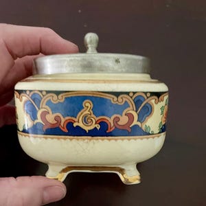 May include: A small, antique ceramic jar with a silver lid. The jar is cream-coloured with a blue and gold decorative band. The jar has a small, round silver knob on top and sits on three small feet.